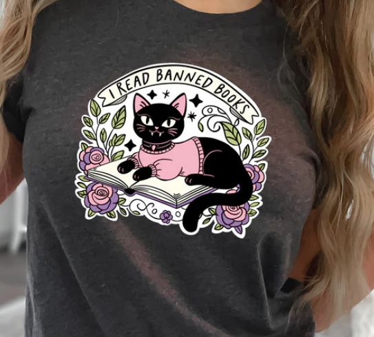 Dark gray t-shirt with black cat in pink sweater lying on an open book surrounded by flowers and leaves, with 'I Read Banned Books' banner above