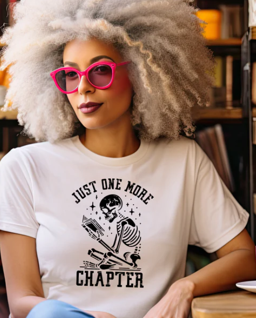 Woman wearing white t-shirt with skeleton reading book graphic and 'Just One More Chapter' text